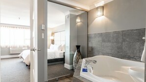 A modern hotel bathroom with a large bathtub, a glass shower enclosure, and a visible bedroom with a bed and a nightstand. - Crowne Plaza Moncton-Downtown by IHG (Moncton)