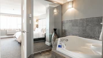 Een moderne hotelbadkamer met een ruime badkuip, een douchecabine van glas en een zichtbare slaapkamer met een bed en nachtkastje.