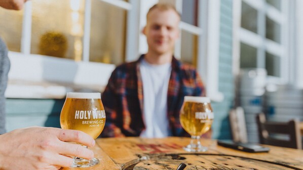 A person holding a glass with 'HOLY WHALE BREWING CO.' text, another glass on the table, and a wooden table in a room with windows.