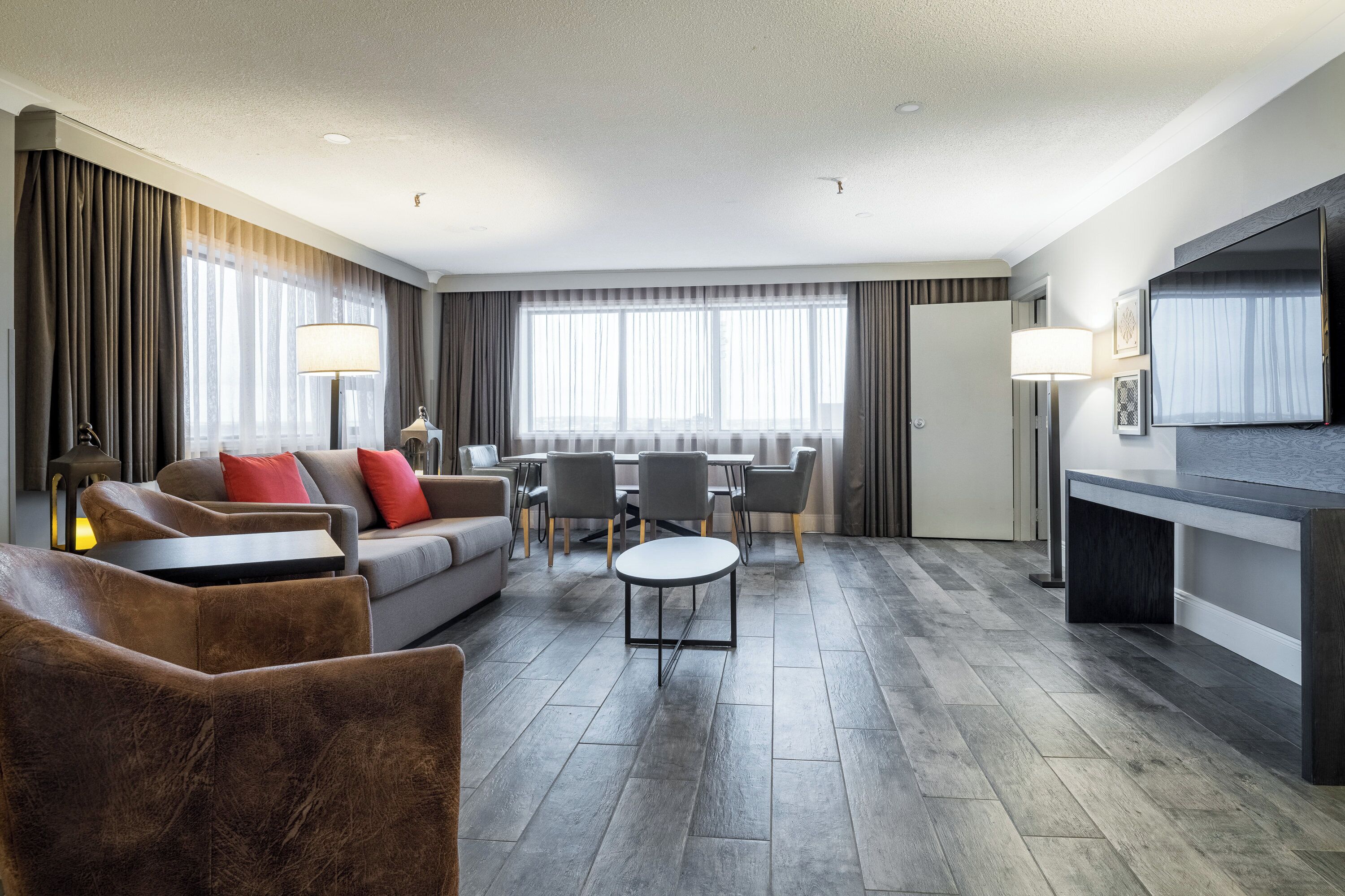 Une chambre d’hôtel moderne équipée d’un canapé, de fauteuils, d’une table à manger, d’un téléviseur et d’une grande fenêtre avec des rideaux.