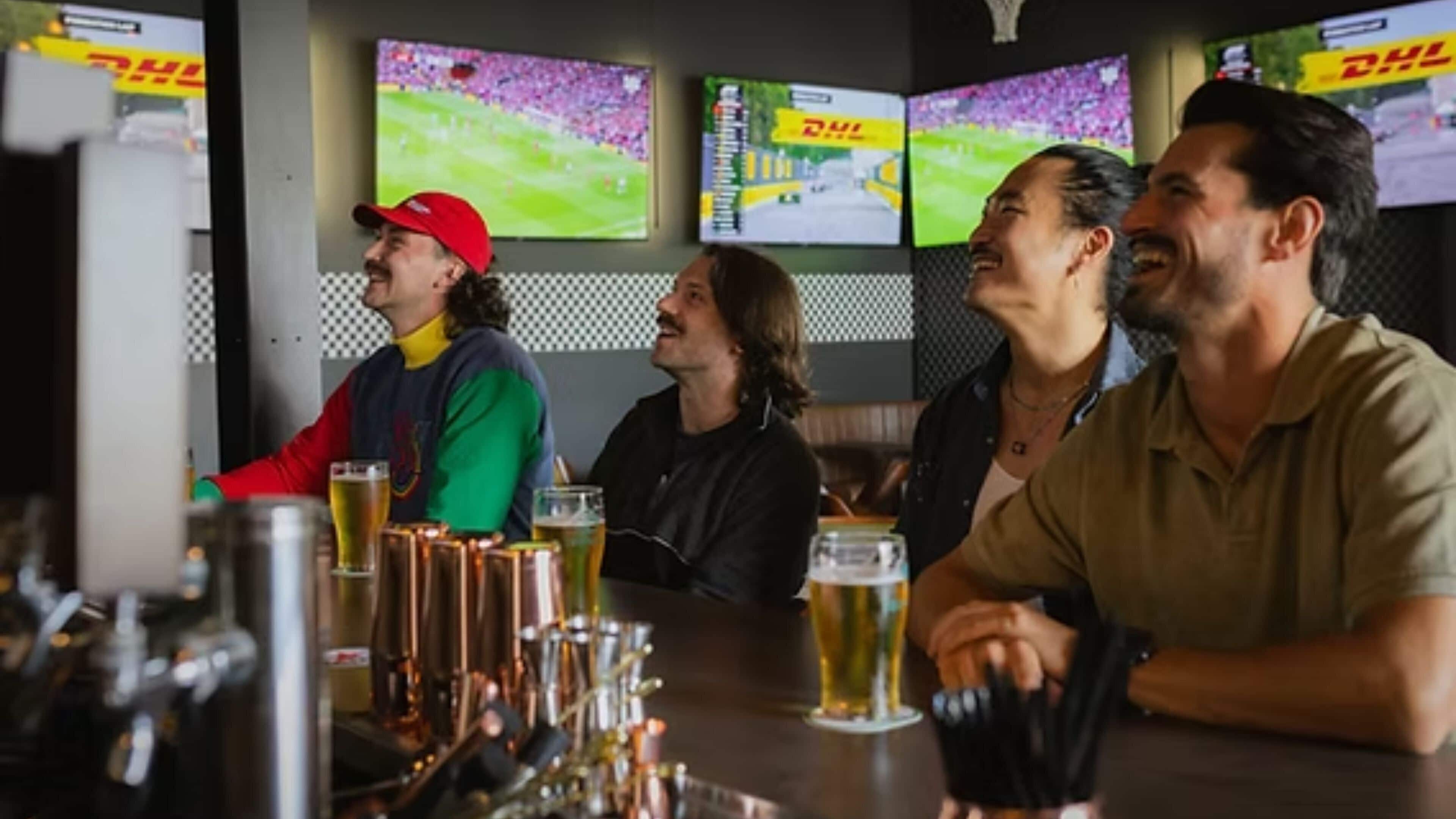 A bar area with multiple screens displaying sports events, a counter with glasses, and patrons seated.