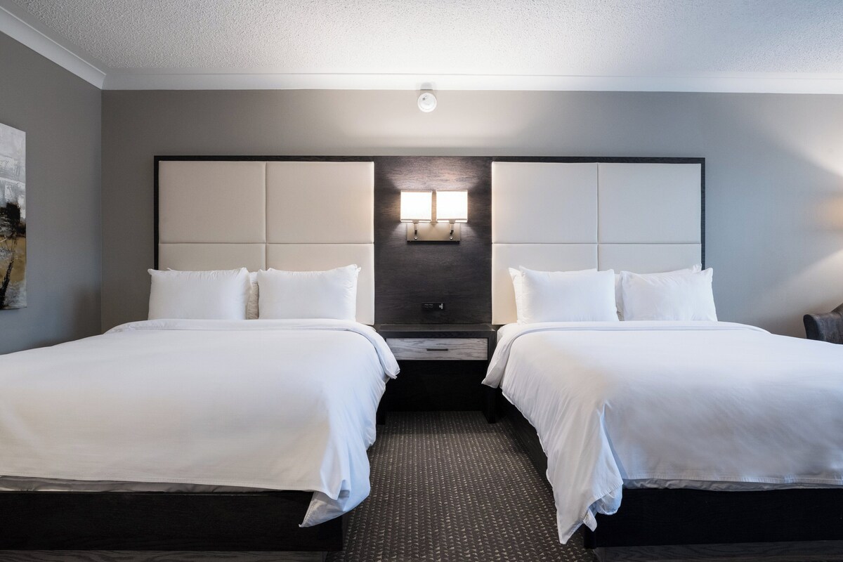 A hotel room with two beds, a dark headboard, a small bedside table, and a wall-mounted light fixture.