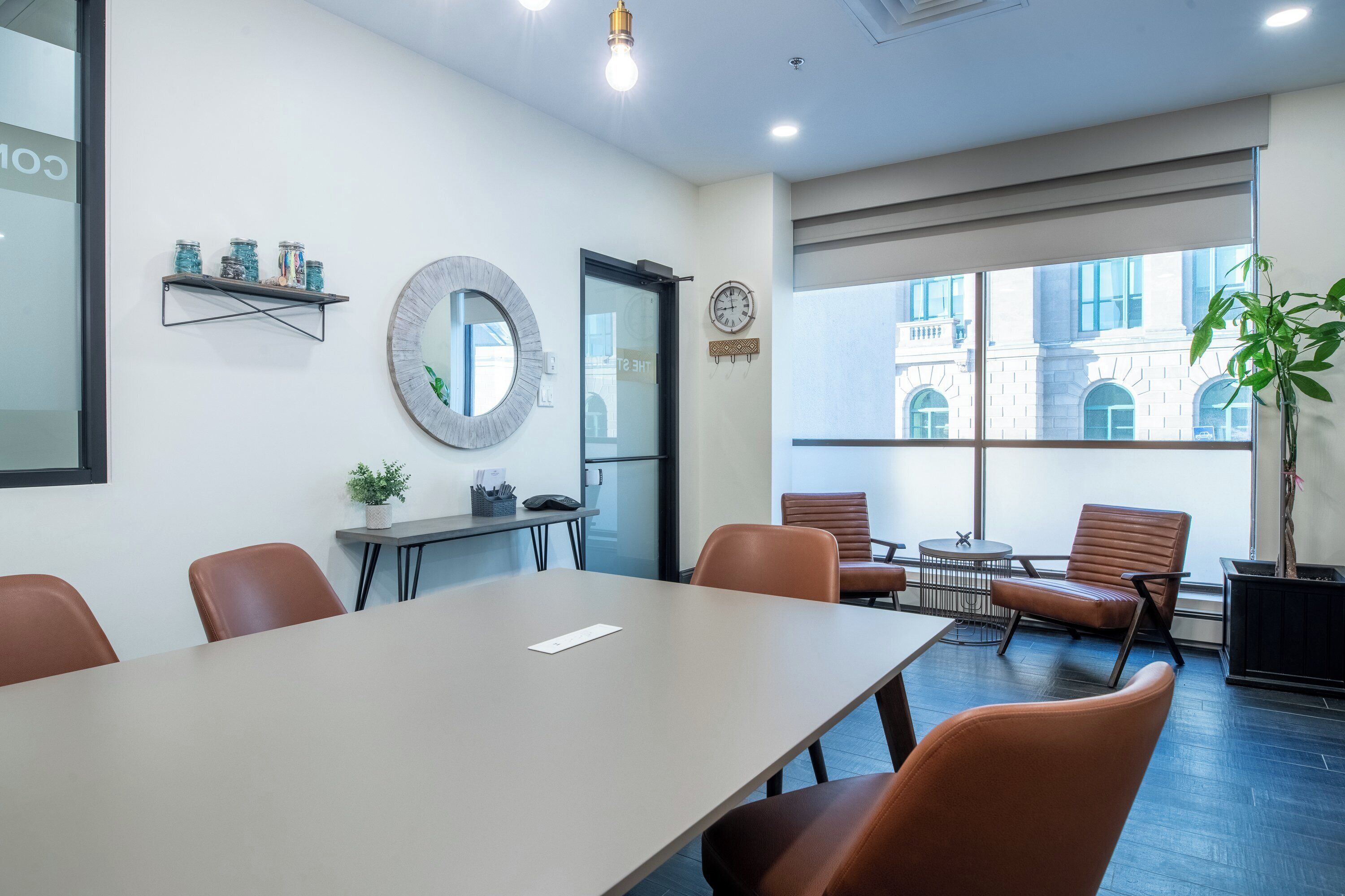 Een vergaderzaal met een lange tafel, diverse stoelen, een klok en een raam met uitzicht op gebouwen.