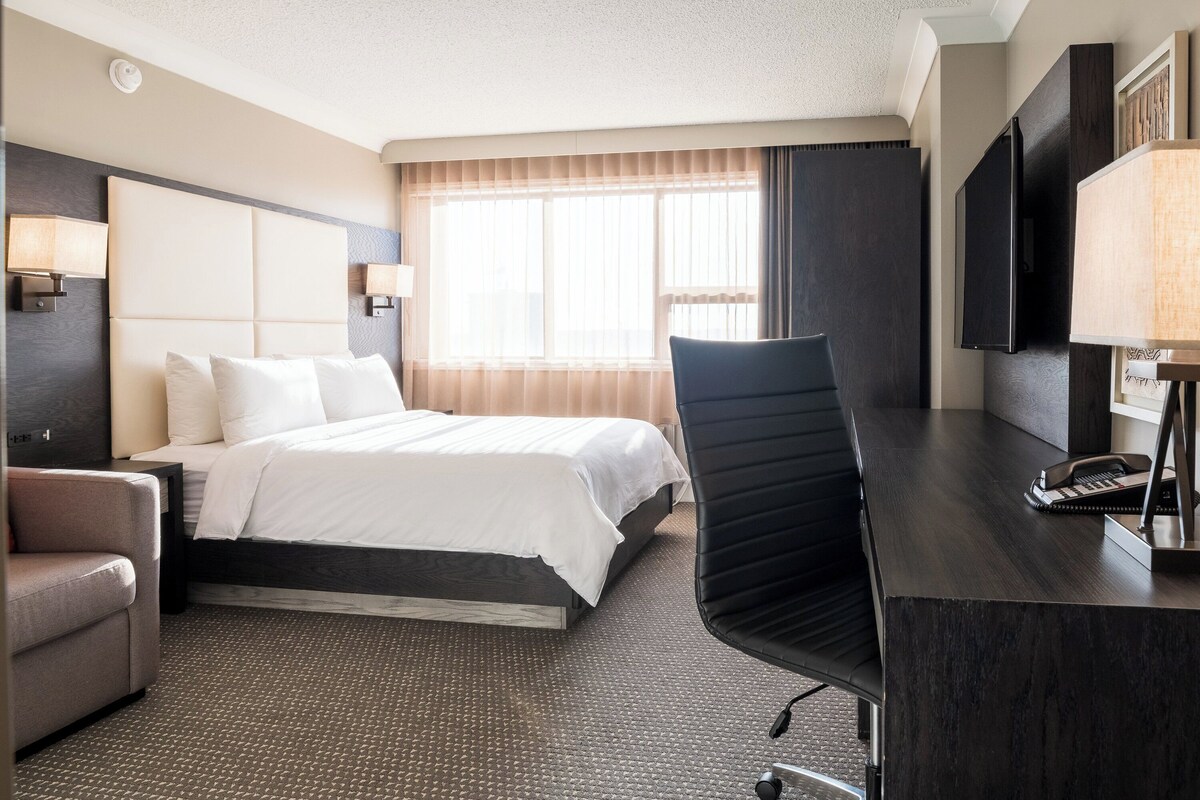 A hotel room with a bed, a desk, a chair, a TV, and a lamp.