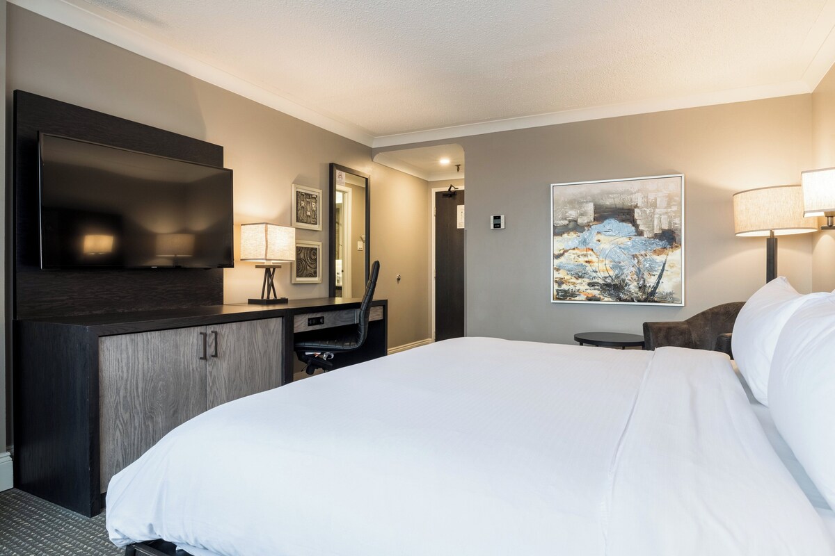 A hotel room with a large bed, a desk with a chair, a flat-screen TV, a painting on the wall, and a lamp.