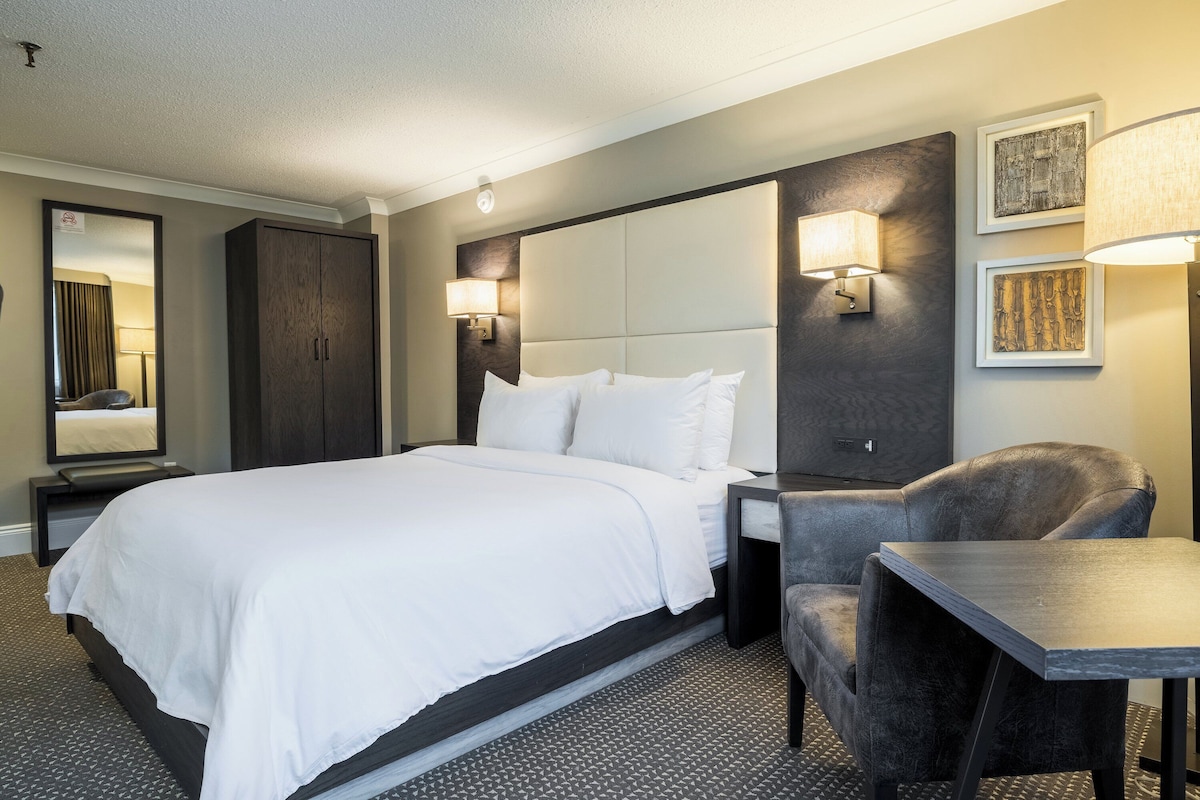A hotel room with a large bed, a desk, a chair, a wardrobe, and wall-mounted lamps.