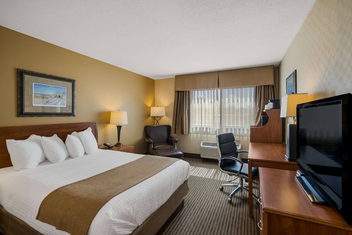 A hotel room with a large bed, a desk with a television, a chair, and a window with curtains.