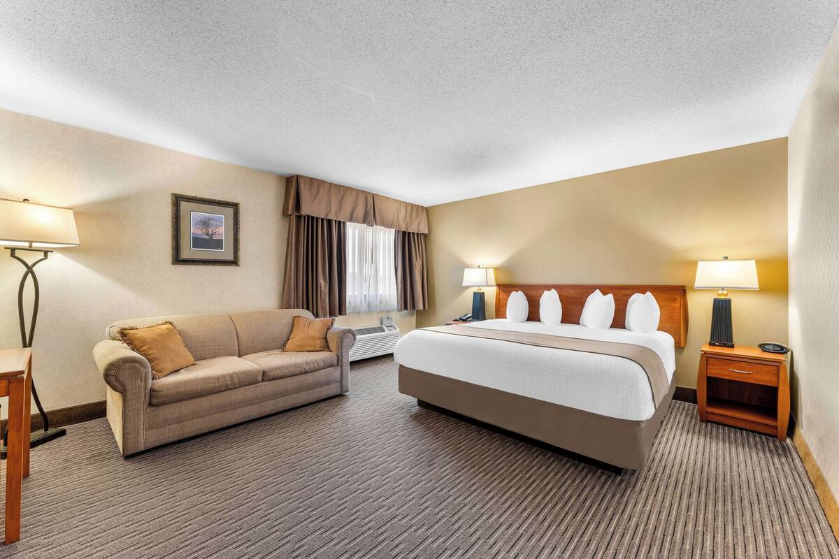 A hotel room with a bed, a sofa, two bedside tables, a lamp, a picture on the wall, and a window with curtains.