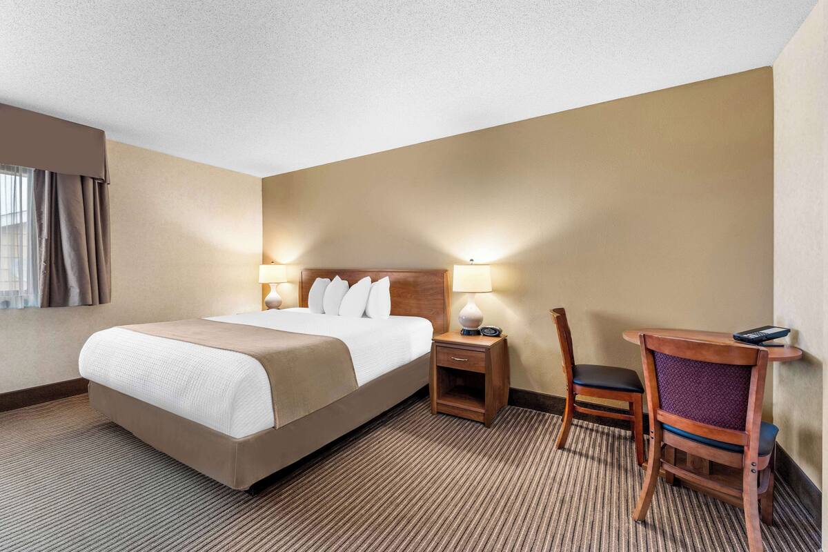 A hotel room with a large bed, bedside lamps, a wooden nightstand, a chair, and a small table with a remote control.