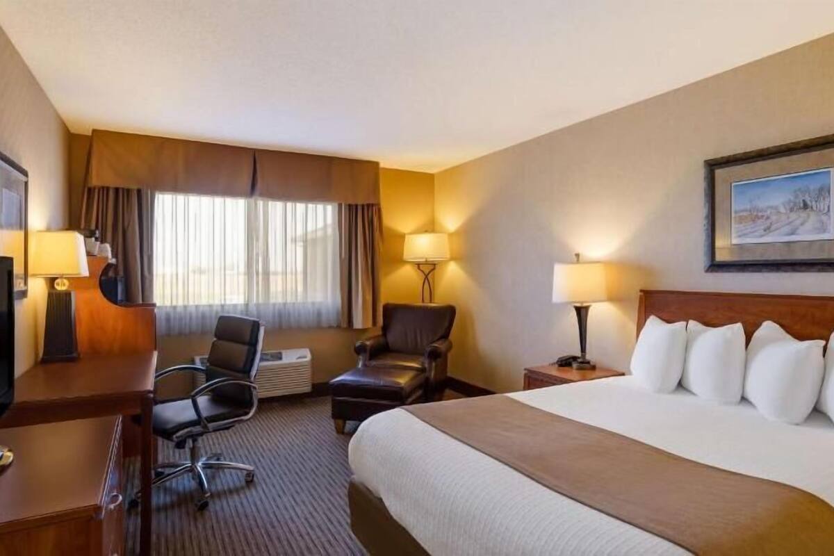 A hotel room with a bed, two chairs, a desk with a TV, a lamp, and a painting on the wall.