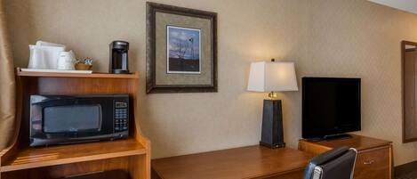 A hotel room with a wooden desk, a microwave, a coffee maker, a television, and a framed picture on the wall.