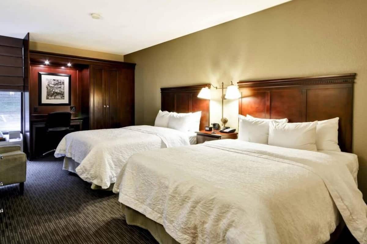 A hotel room with two beds, a desk, a chair, a lamp, and a framed picture on the wall.
