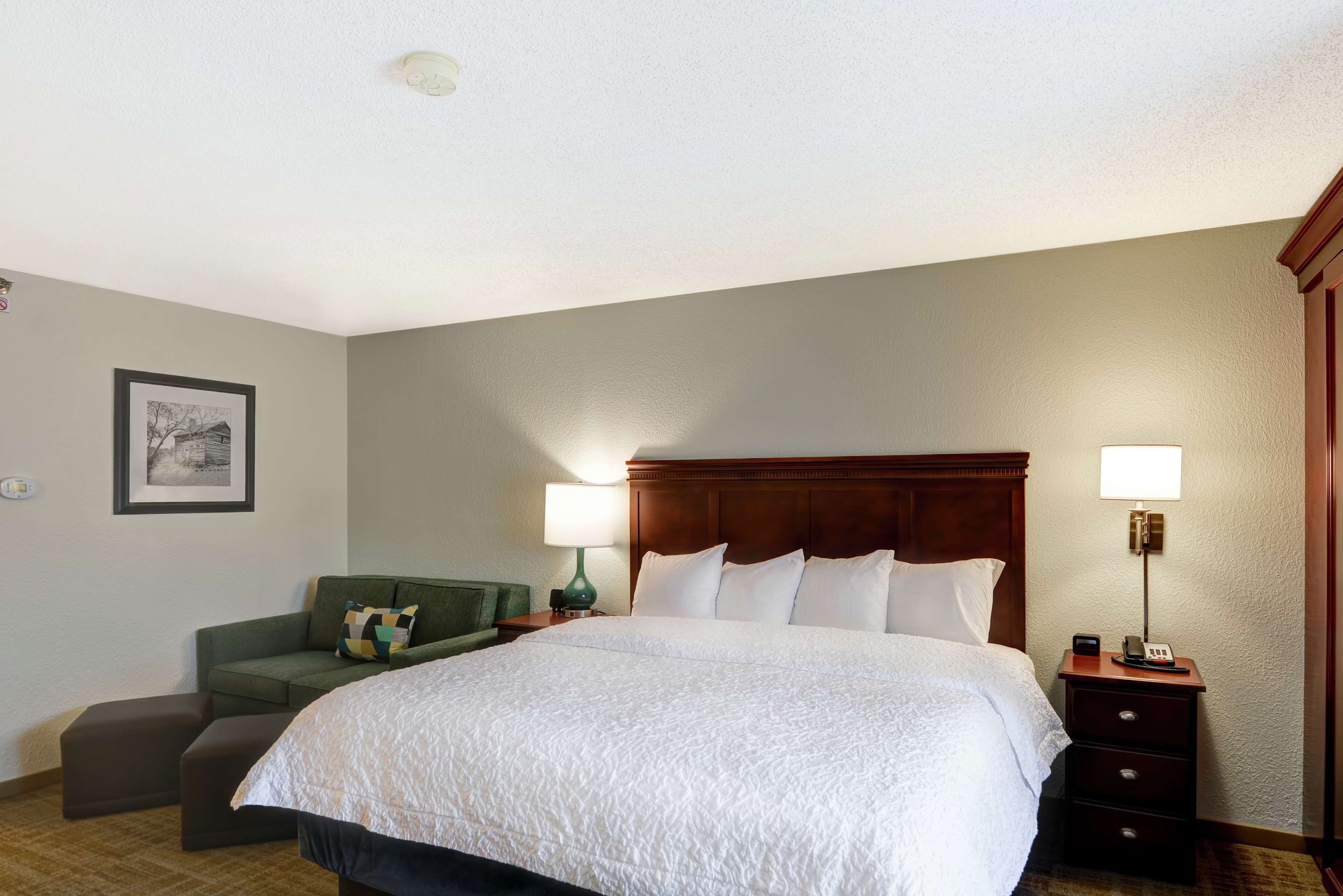 A neatly arranged hotel room with a large bed, a green sofa, a nightstand with a lamp, and a framed picture on the wall.