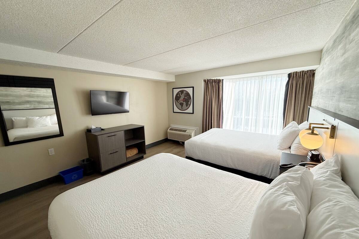 Ein Hotelzimmer mit zwei Betten, einem Flachbildfernseher, einem Schreibtisch mit Lampe und einem großen Fenster mit Vorhängen.