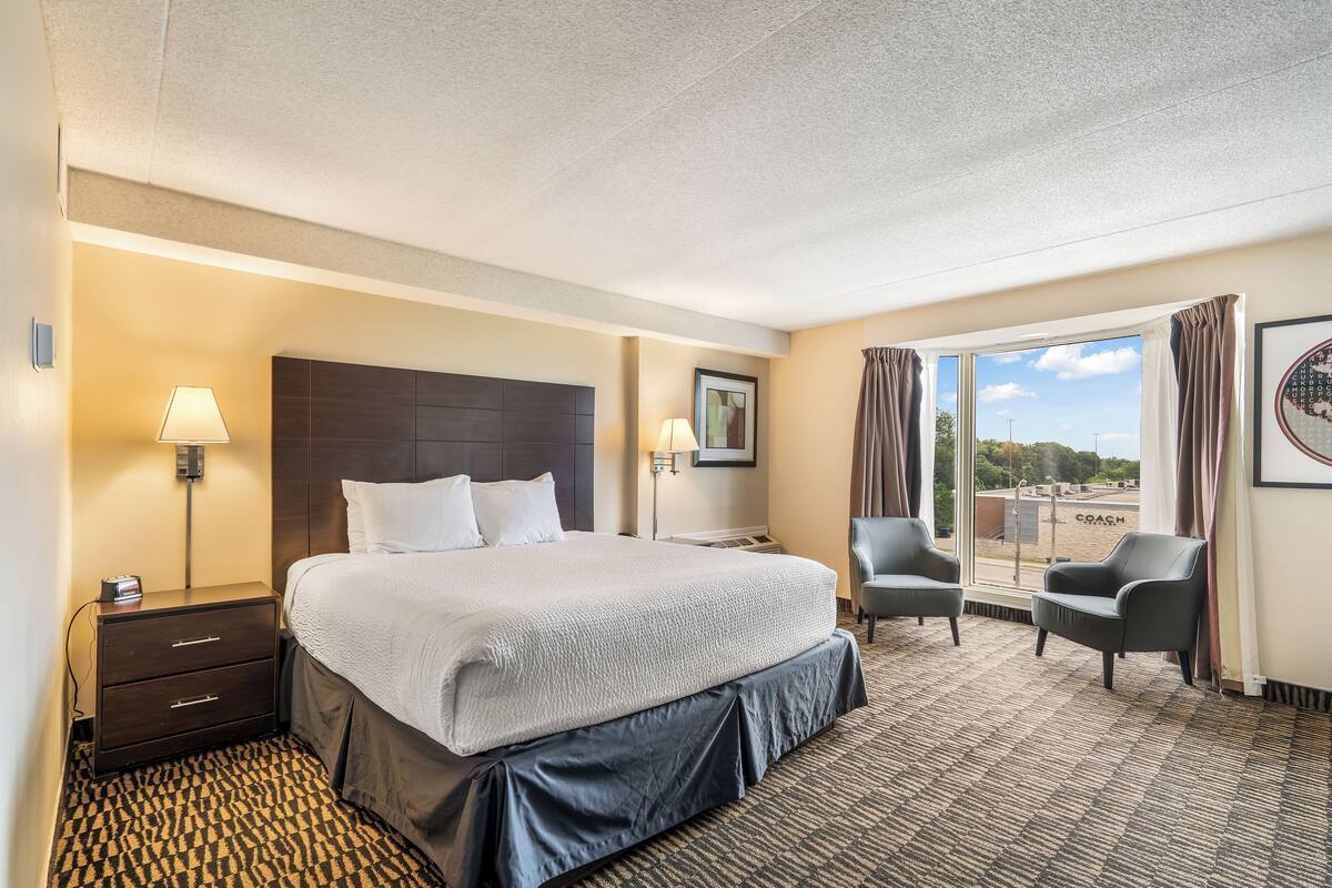 Ein Hotelzimmer mit einem großen Bett, zwei Nachttischen, einem Sessel, einem Fenster mit Blick und einem Teppichboden.