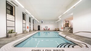 Una piscina interior con una zona poco profunda señalada en el suelo, rodeada de paredes de azulejos y un techo con luminarias lineales.