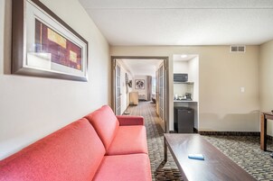 Quarto de hotel com sofá vermelho, mesa de madeira, área de cozinha com micro-ondas e piso estampado.