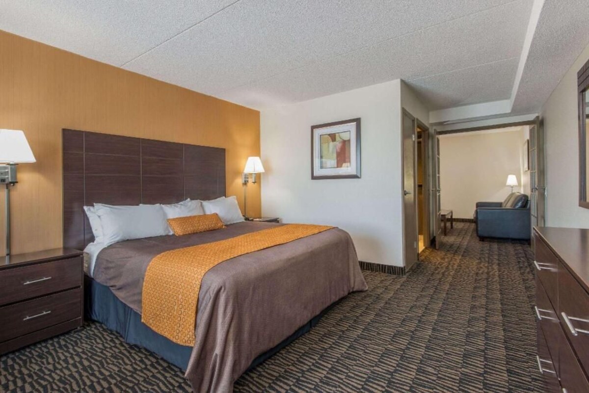 Ein Hotelzimmer mit einem großen Bett, Nachttischlampen, einem Kopfteil, einem Teppichboden, einem Schreibtisch mit Schubladen und einer Tür zu einem anderen Raum.