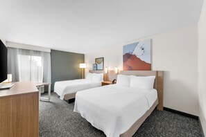 Hotellihuone, jossa on kaksi sänkyä, työpöytä, tuoli, lamppu ja taulu seinällä.