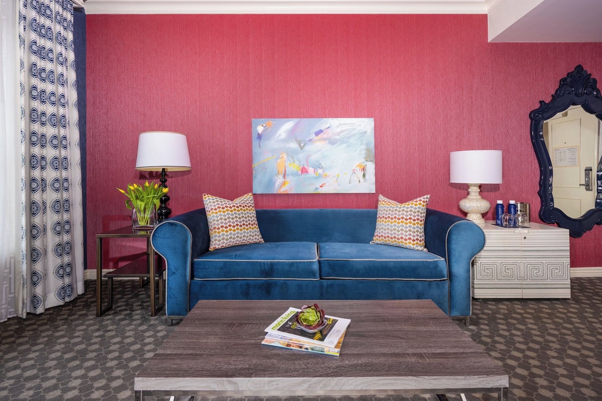 A living room with a blue sofa, two lamps, a coffee table with a magazine, a painting on the wall, and a patterned curtain.