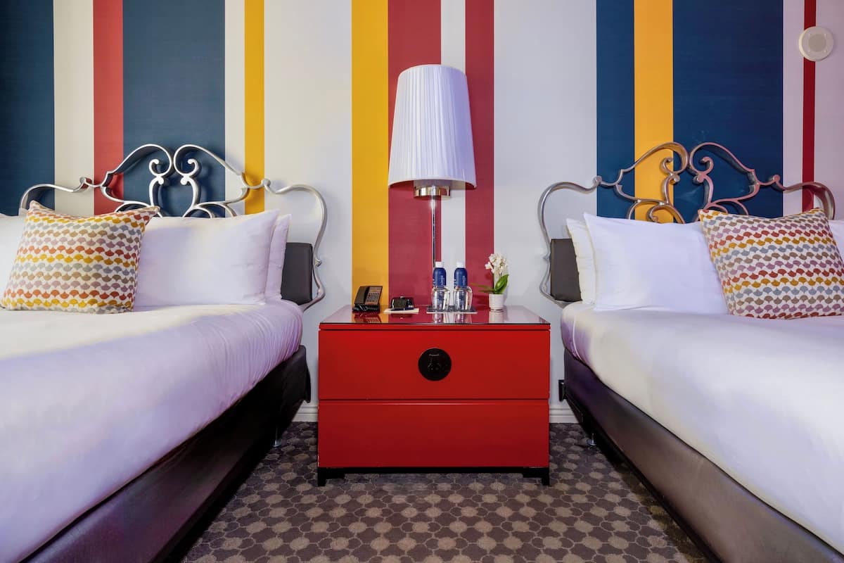 A hotel room with two beds, a red nightstand, a lamp, and a striped wall.