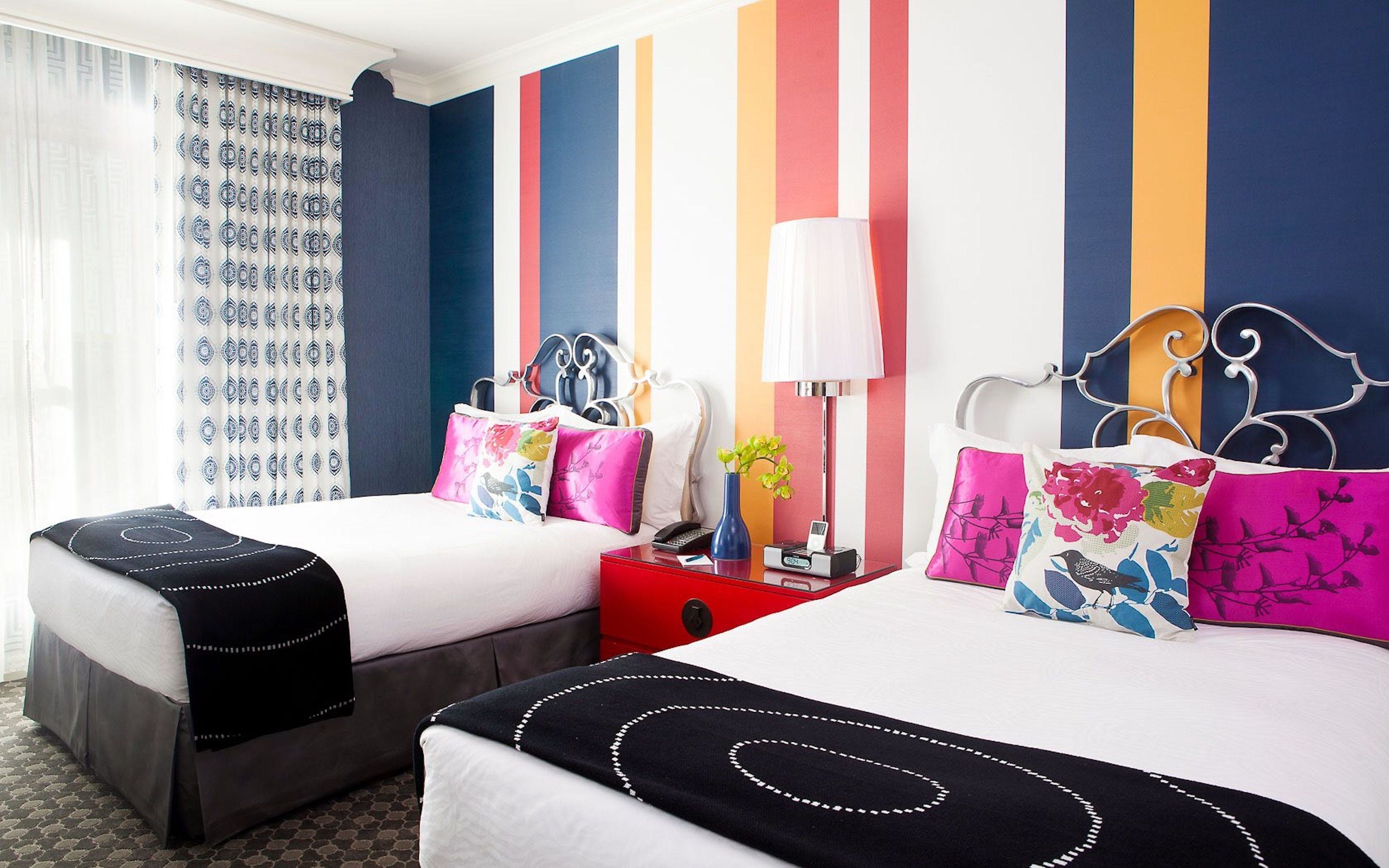 A hotel room with two beds, striped walls, a window with patterned curtains, and a bedside table with a lamp and phone.