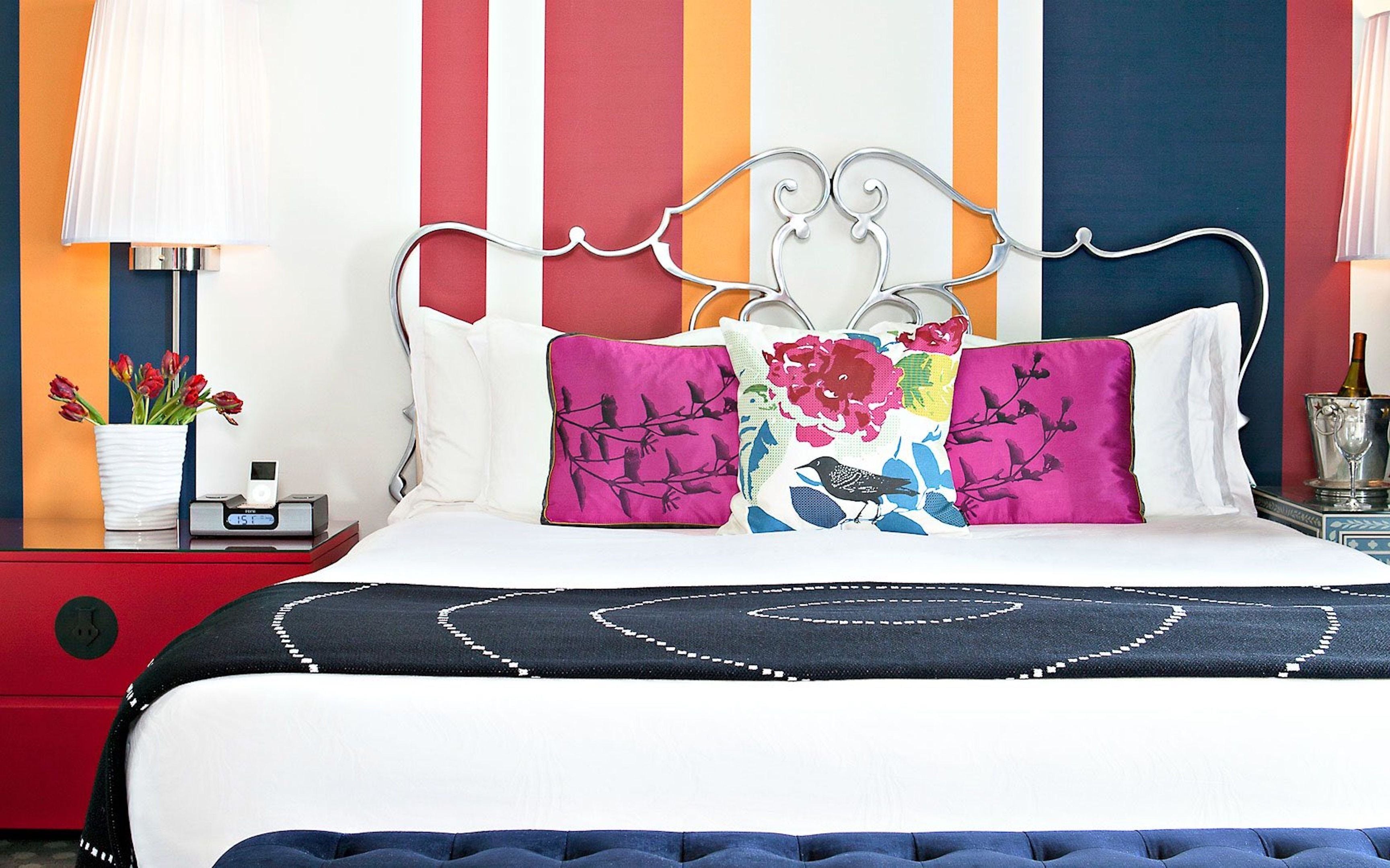 A bed with a metal frame, multiple pillows, a bedside table with a lamp, and a vase with flowers.