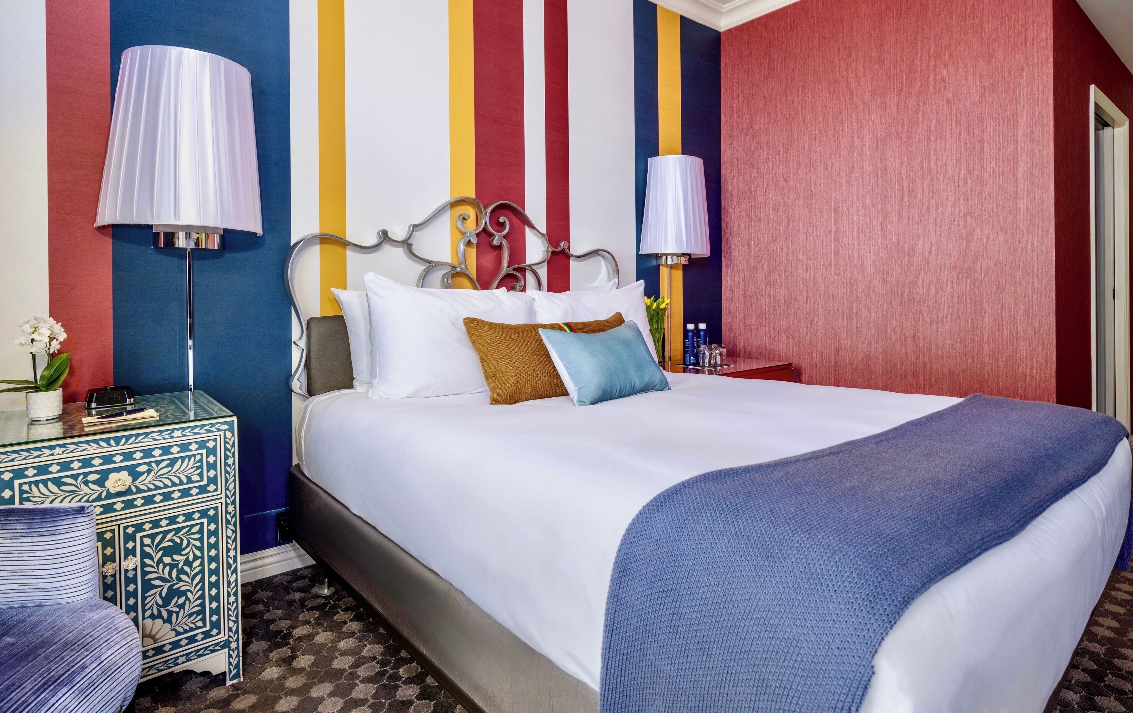 Una cama bien hecha con sábanas blancas, una manta azul y múltiples almohadas de varios colores. Hay una mesita de noche con una lámpara y una planta pequeña. La habitación presenta un diseño de pared a rayas y un cabecero moderno.