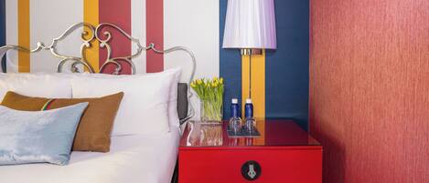 A bed with white bedding and a red nightstand with a lamp and toiletries.