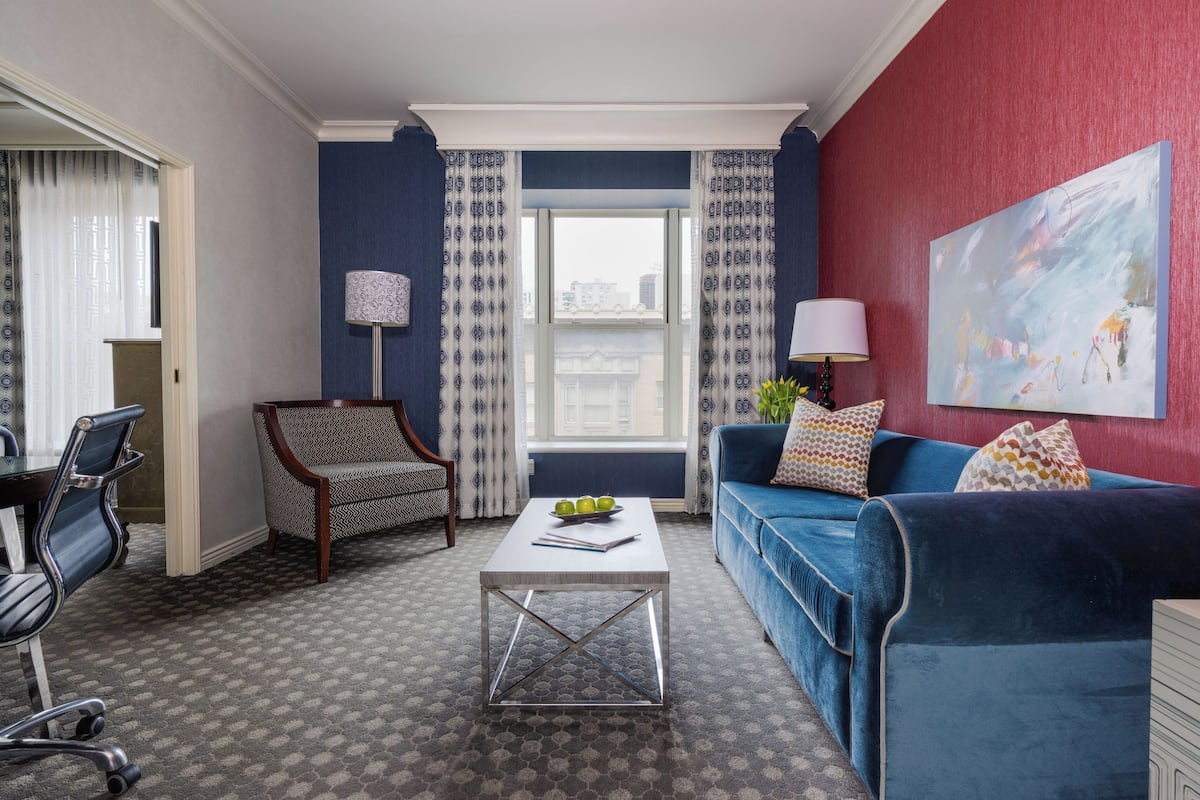 A modern living room with a blue sofa, a patterned armchair, a coffee table with apples, and a large abstract painting on the wall.