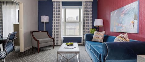 Un salón moderno con un sofá azul, un sillón estampado, una mesa baja con manzanas y un cuadro abstracto grande en la pared.