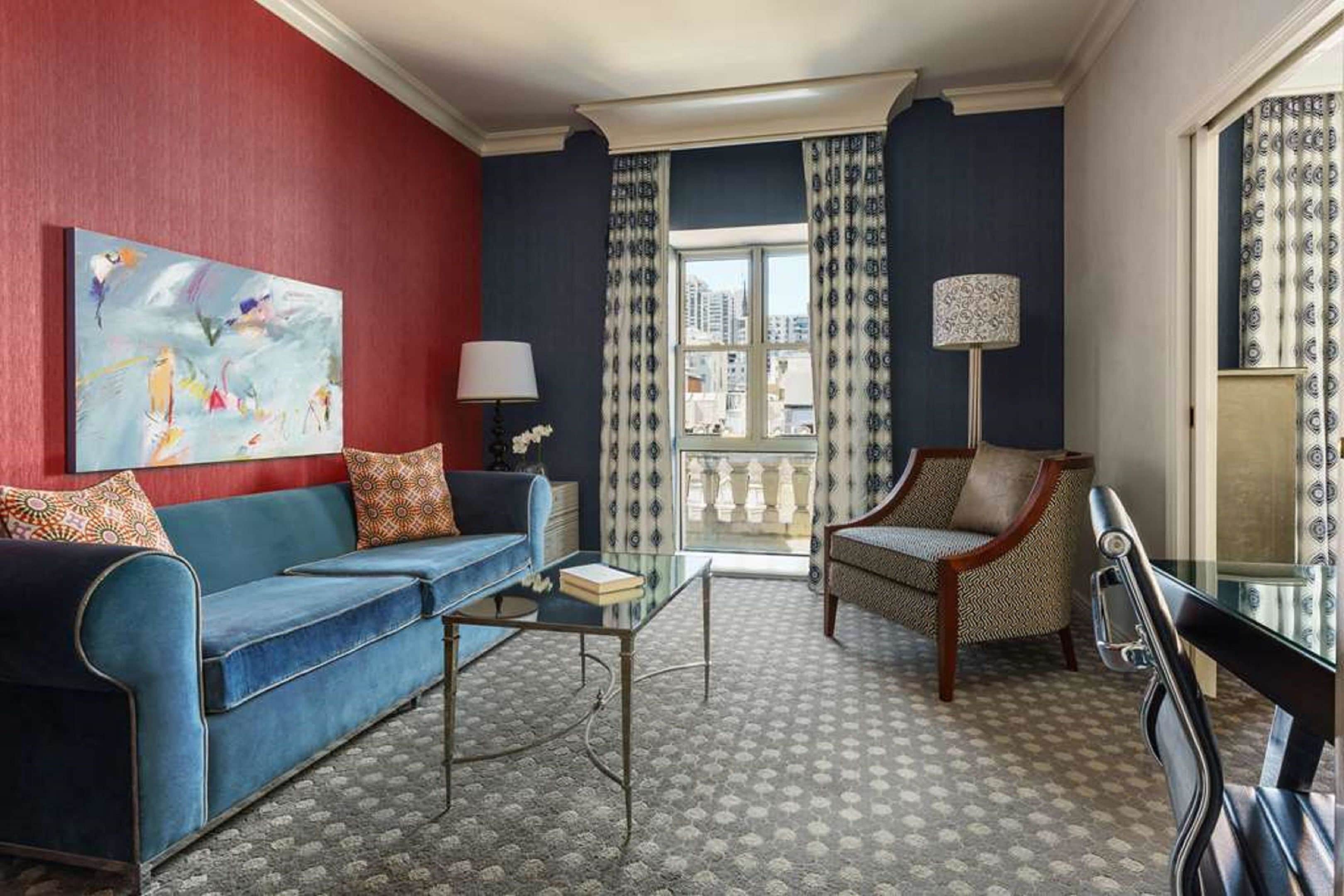 Una sala de estar con un sofá azul, un sillón estampado, una mesa de centro de vidrio y un cuadro en la pared.