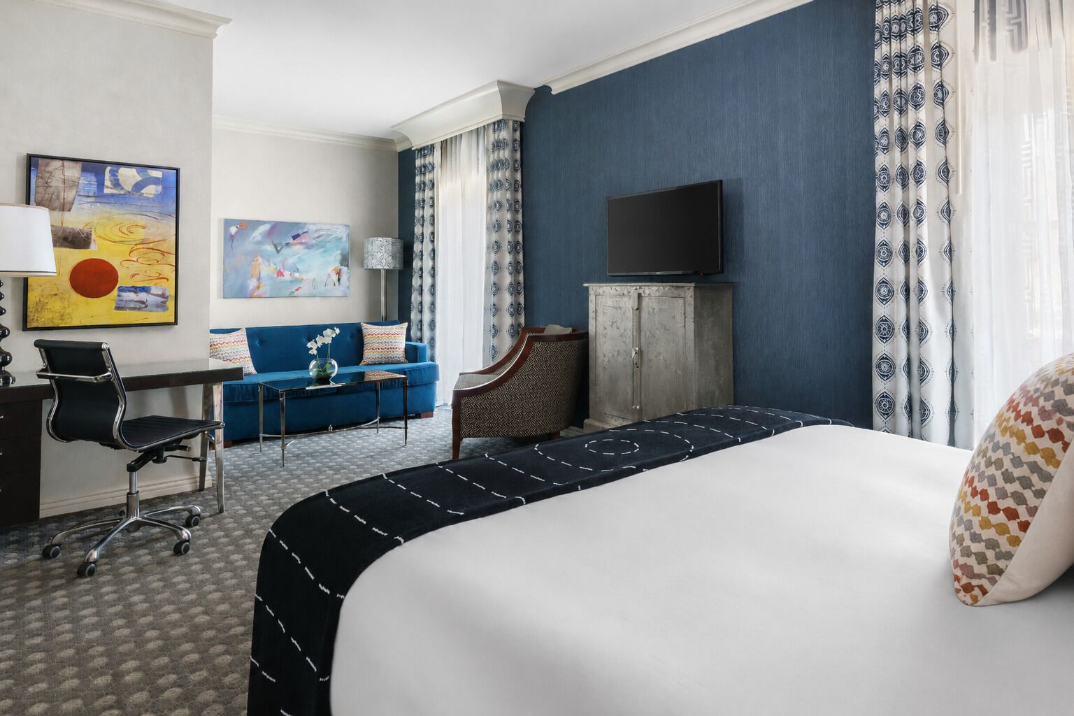 A hotel room with a bed, a desk, a chair, a television, and a painting on the wall.