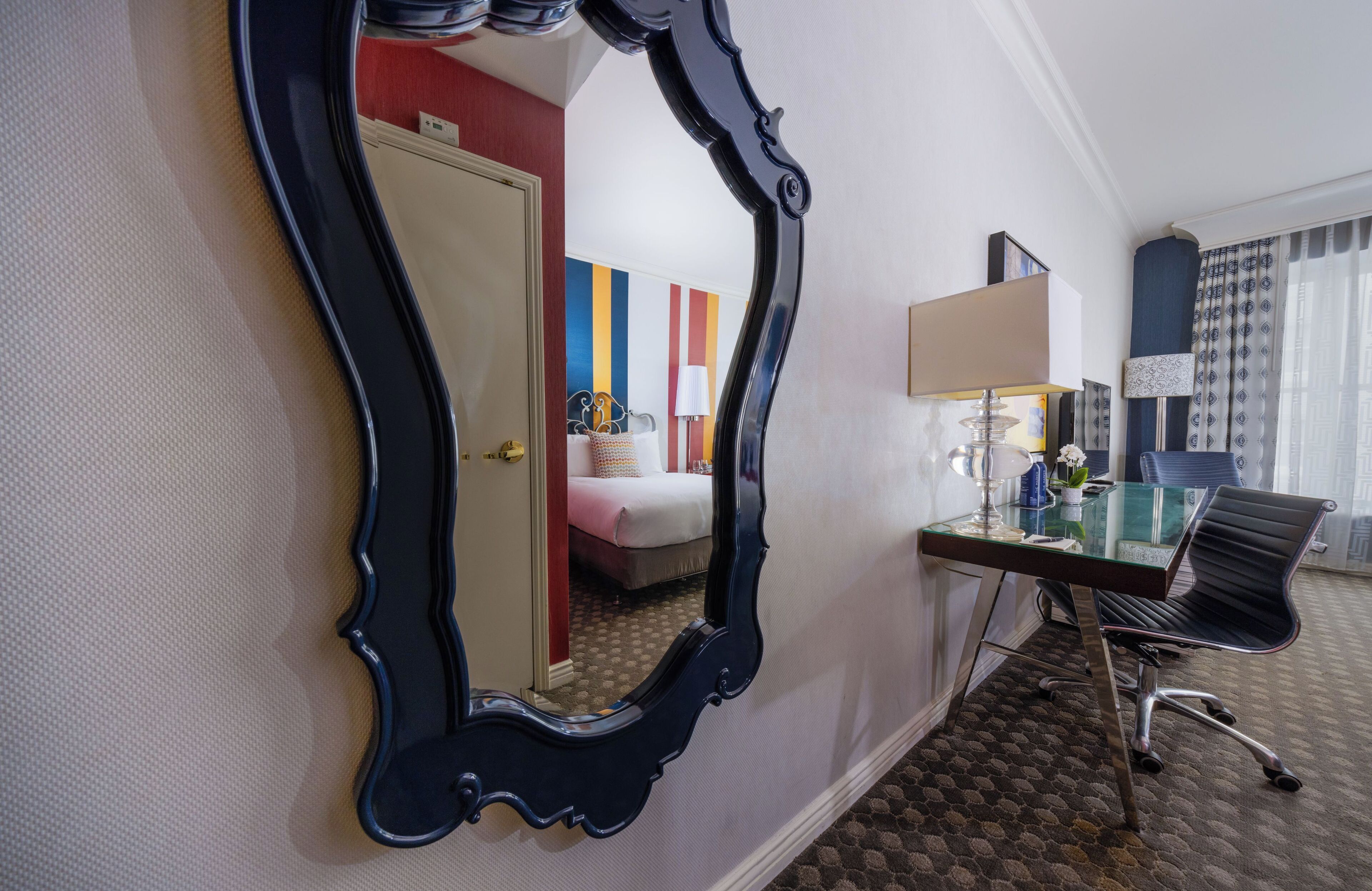 A hotel room with a large mirror, a desk with a lamp, a chair, and a bed with colorful curtains.