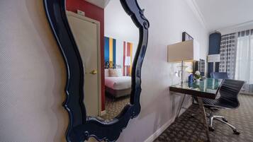 A hotel room with a large mirror, a desk with a lamp, a chair, and a bed with colorful curtains.
