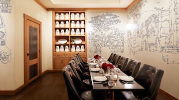 A dining area with a long table set for four, surrounded by black leather chairs. A wooden cabinet with white and blue patterned vases is against the wall.