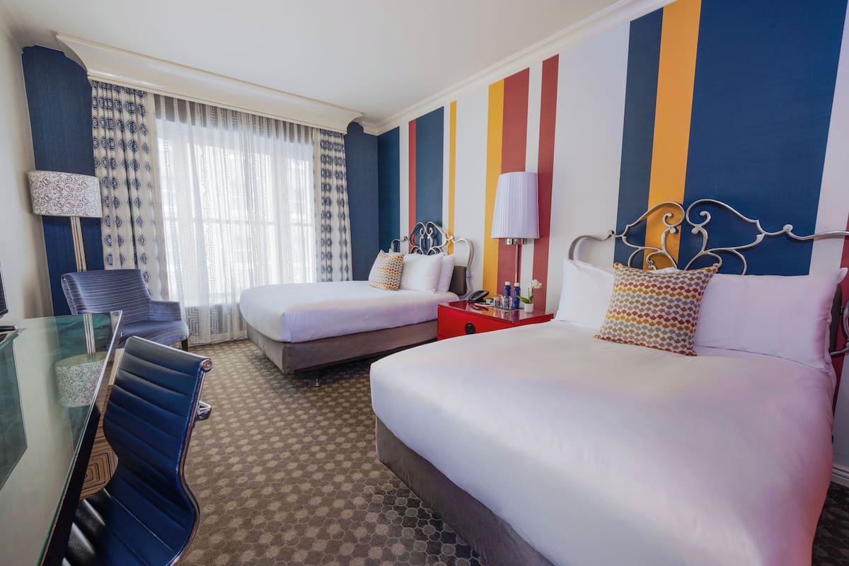 A hotel room with a glass table, two beds, a red nightstand, and a patterned wall.