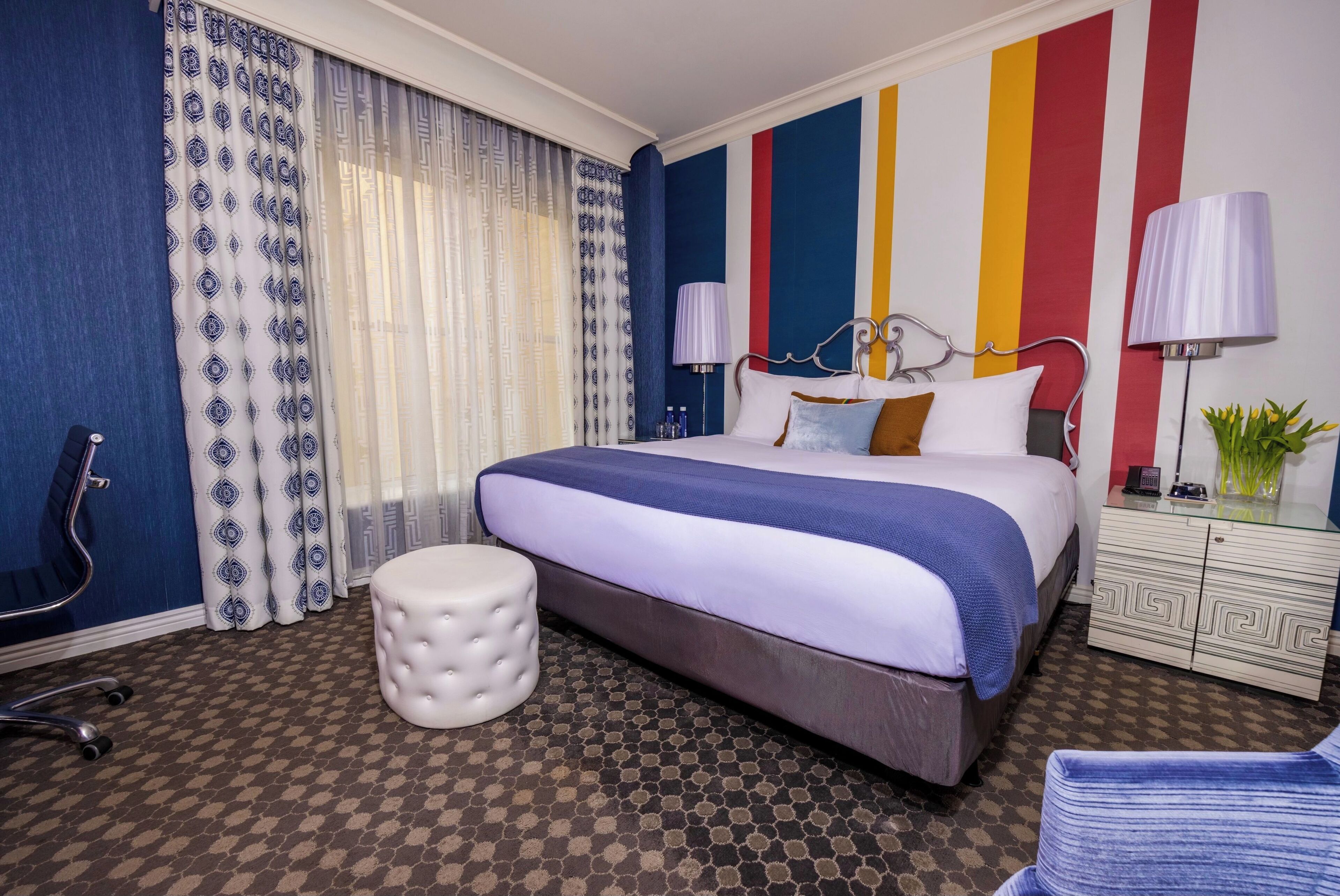 A hotel room with a large bed, a white ottoman, a desk with a lamp, and a wall with vertical stripes.