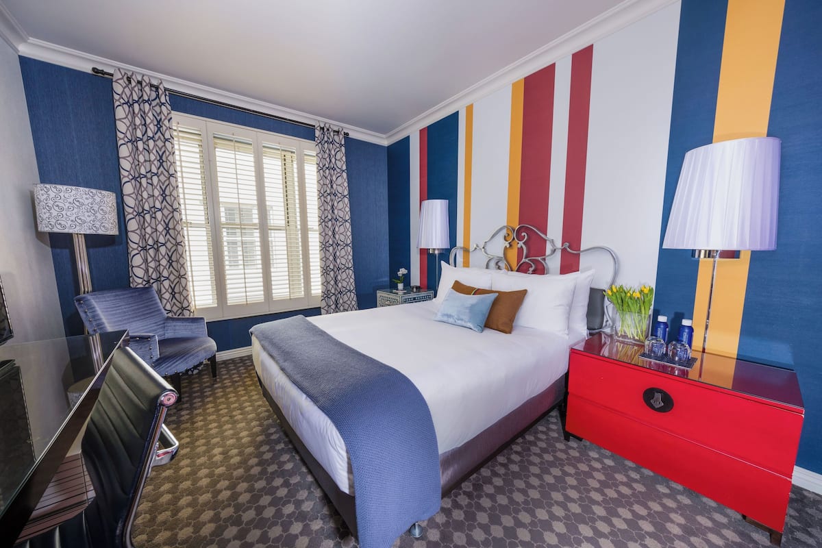 A hotel room with a bed, a desk, a chair, a nightstand, and a lamp.