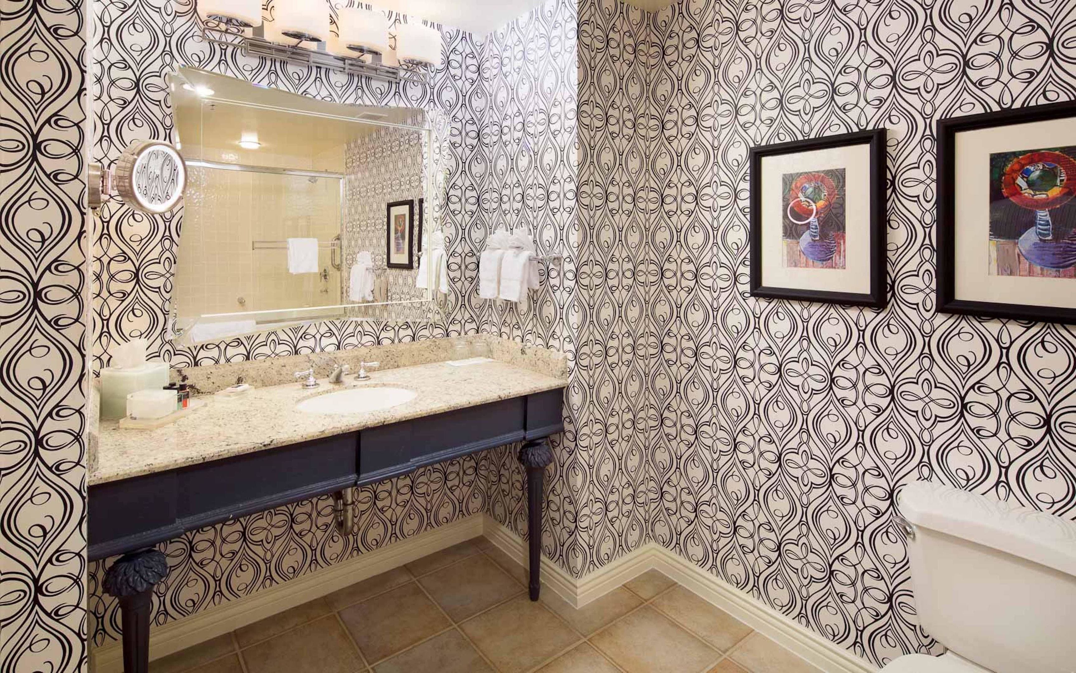 A bathroom with a patterned wallpaper, a double sink vanity, and framed artwork on the wall.