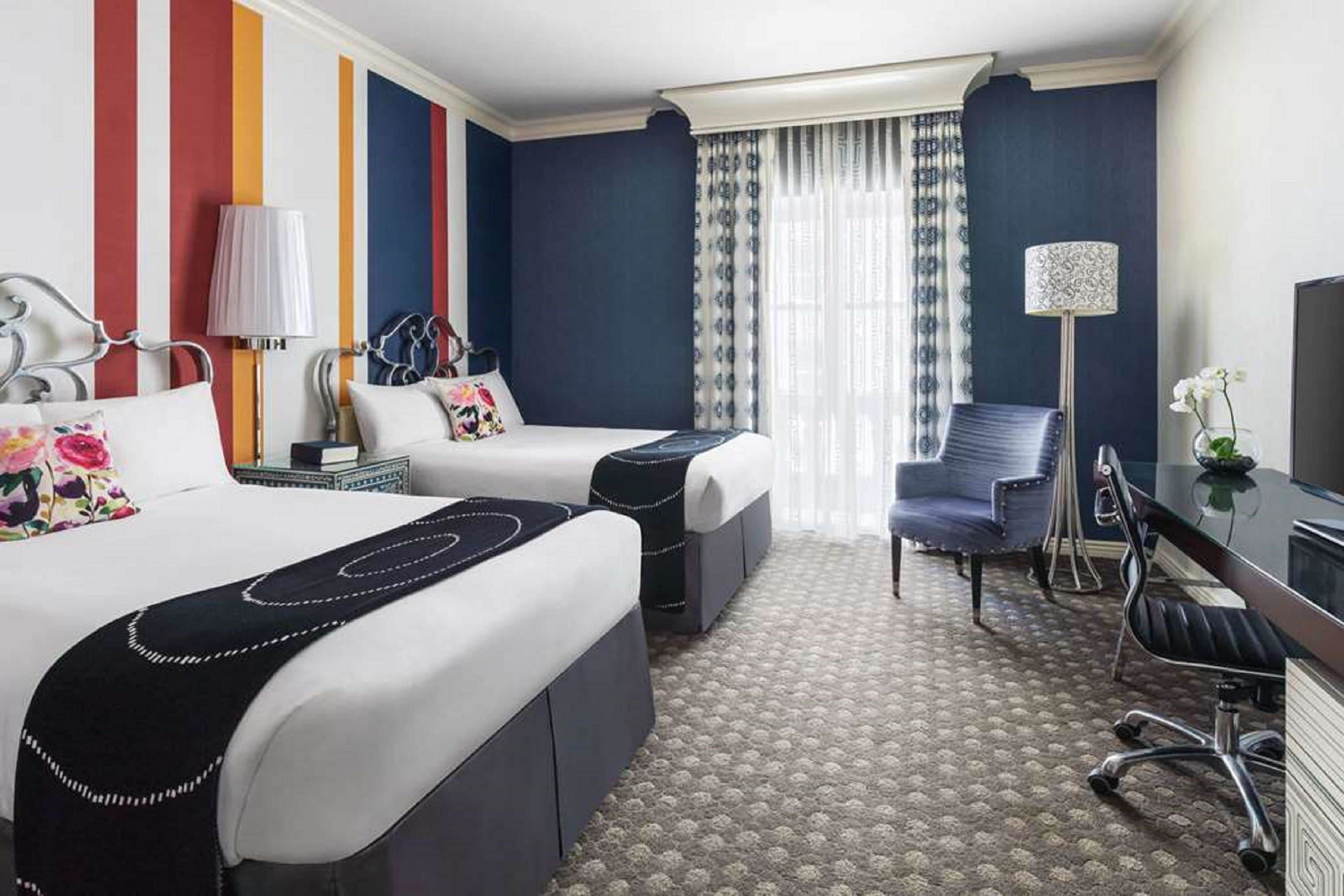 A hotel room with two beds, a desk, a chair, a TV, and a window with curtains.