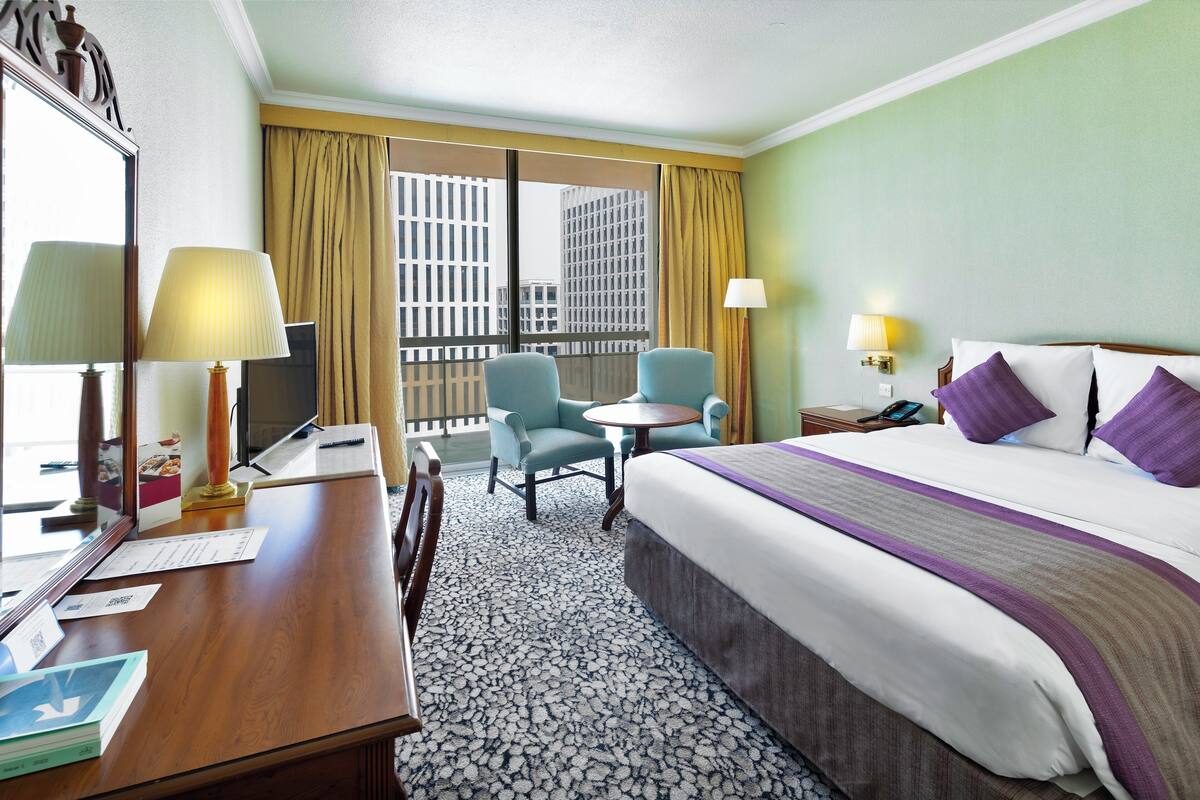 Habitación de hotel con cama, dos sillas, escritorio, lámpara, mesita de noche y ventana con cortinas.