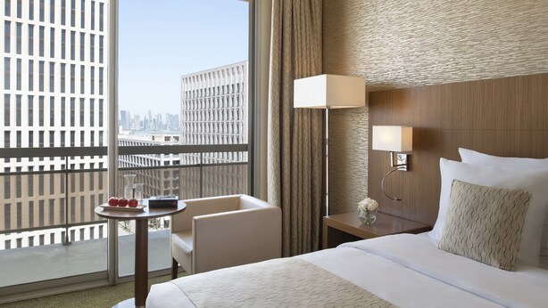 Una habitación de hotel con un ventanal grande, una cama con ropa blanca, una mesita de noche con lámpara y vistas a un paisaje urbano.