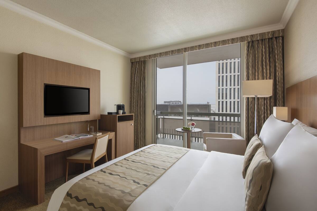 Habitación de hotel con cama, escritorio con televisor de pantalla plana, silla, mesita de noche con lámpara y balcón con vistas a la ciudad.