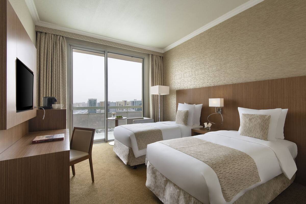 Habitación de hotel con dos camas, televisor, escritorio y un ventanal con vistas a la ciudad.