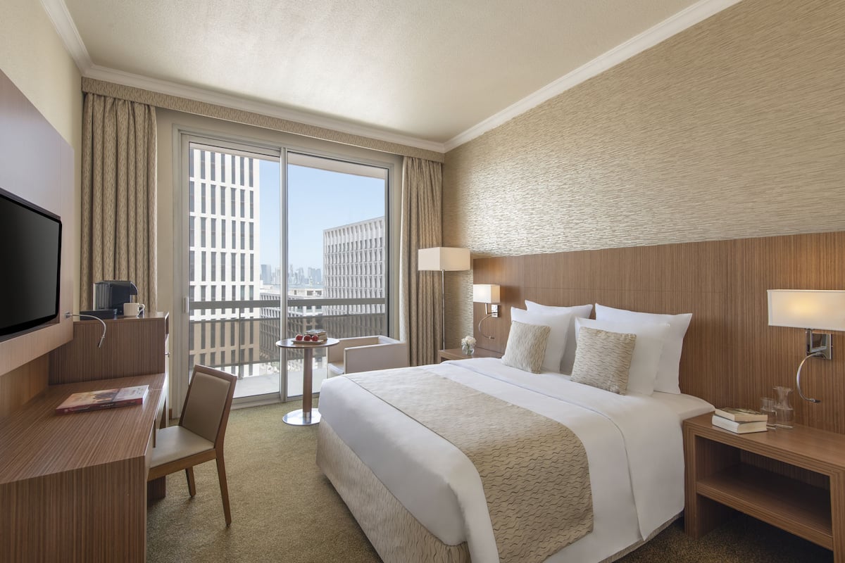 Habitación de hotel con una cama grande, un escritorio con televisor de pantalla plana, una silla, una mesita con una bebida y vistas de la ciudad a través de amplios ventanales.