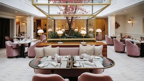 A spacious hotel dining area with pink upholstered seating, a central table with a glass top, and a large decorative tree in the background.