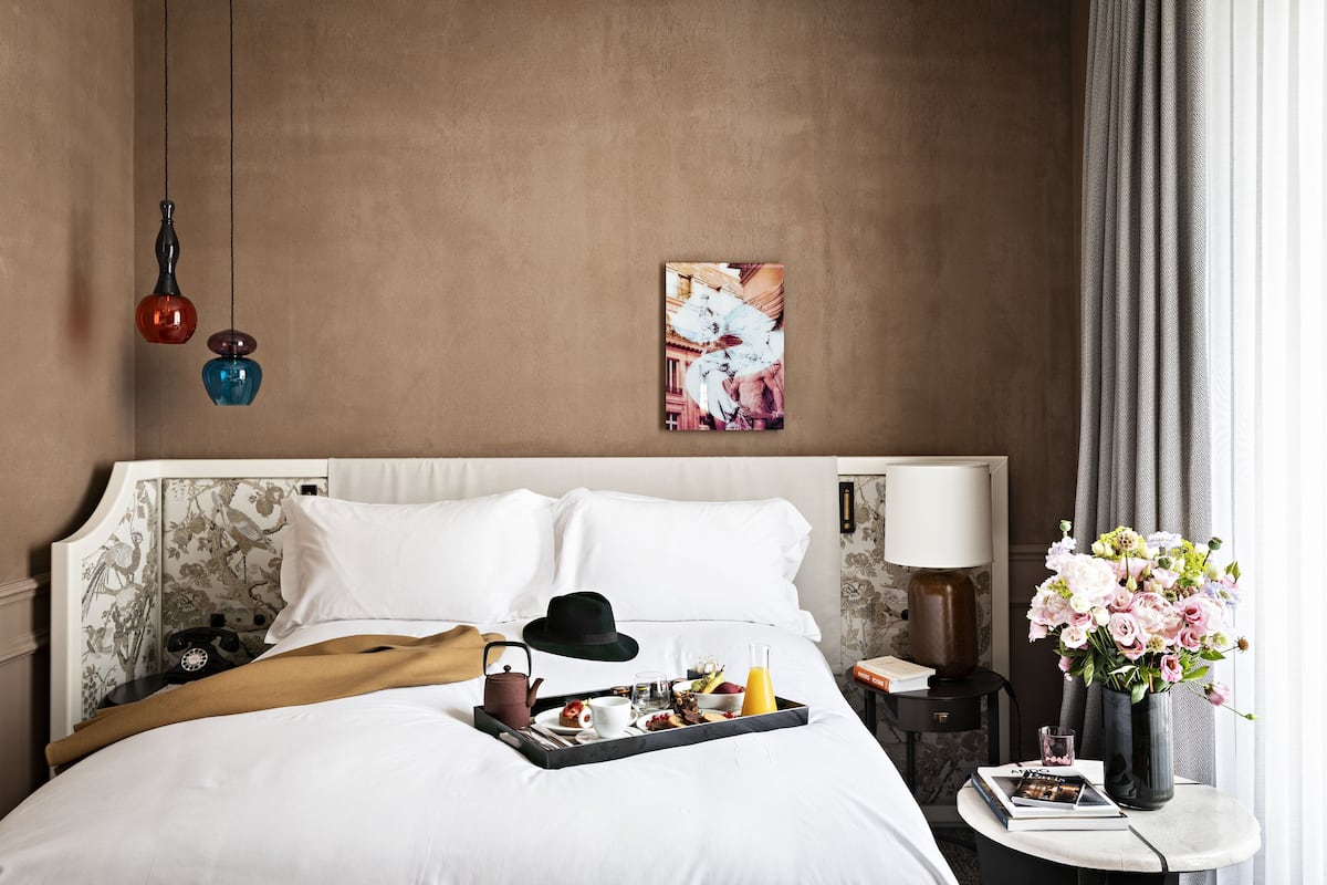 A neatly made bed with white linens, a tray with breakfast items, a black hat, and a vase with flowers on a bedside table.