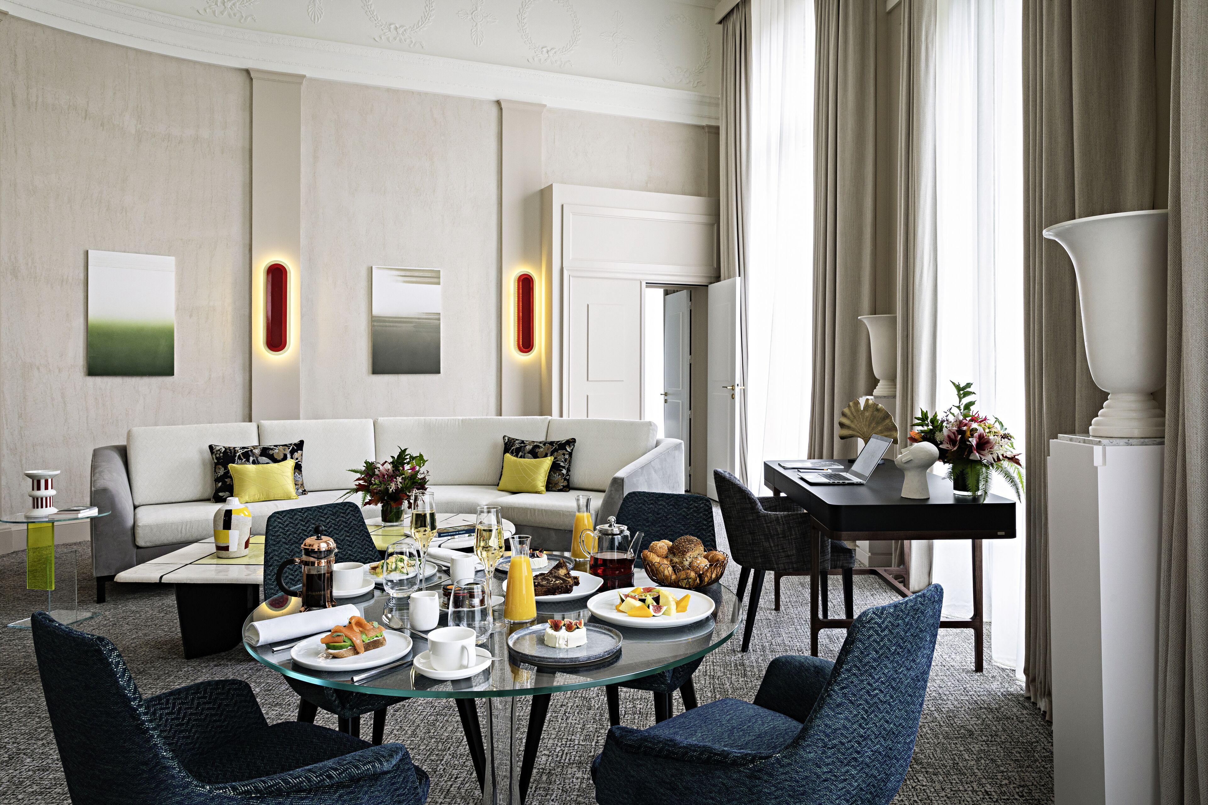 A modern hotel room with a dining area, a sofa set, and a dining table with breakfast items.