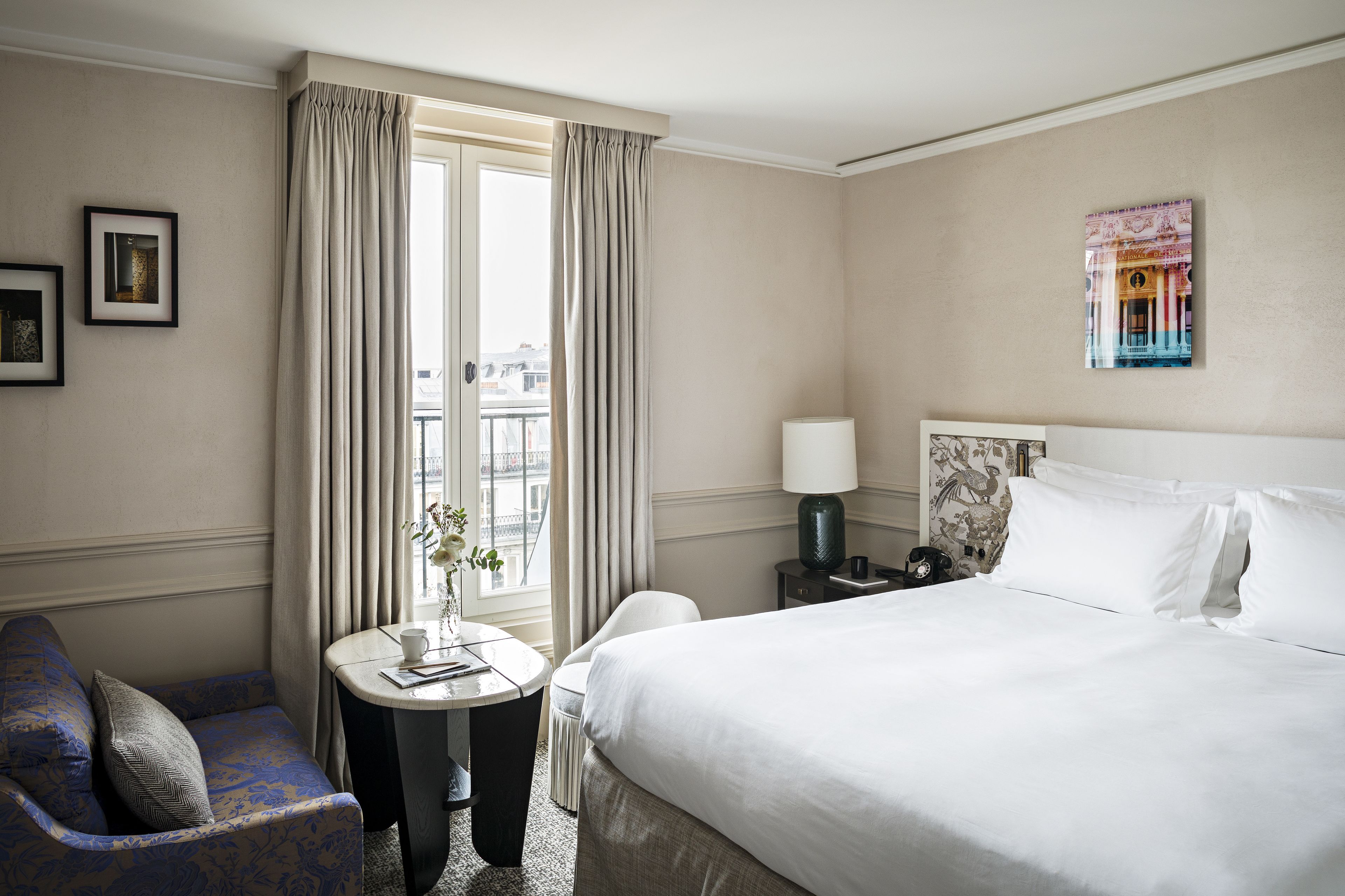 A hotel room with a large bed, a small round table with a lamp, a sofa, and a window with curtains.